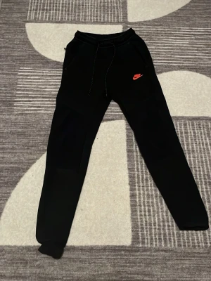 Äkta Nike tech byxor använd lite  - Svarta mjukisbyxor från Nike med röd logga på vänster sida. Byxorna har dragsko i midjan och smal passform med muddar vid bensluten. Perfekta för chill eller träning. Materialet är mjukt och bekvämt.