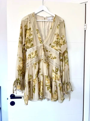 Blommig kortklänning med volang - Romantisk kortklänning i beige med grönt och gult blommönster. Klänningen har v-ringning, spetsdetaljer, volanger och knytband framtill samt långa ärmar