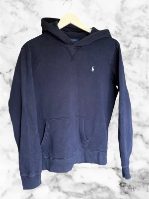 Mörkblå hoodie från Polo Ralph Lauren - Klassisk mörkblå hoodie från Polo Ralph Lauren med huva och känguruficka framtill. Diskret broderad logga på bröstet. Tillverkad i mjukt bomullsmaterial och har ribbade muddar vid ärmslut och nederkant. Perfekt för en avslappnad och stilren look.