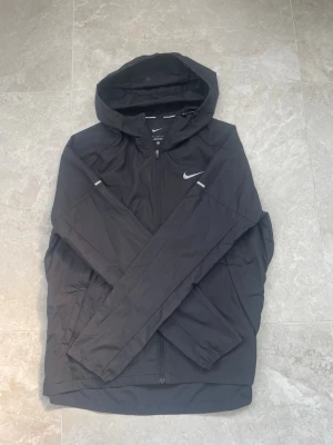 Svart Nike Miller - Snygg svart vindjacka från Nike med huva och dragkedja framtill. Jackan har reflekterande detaljer på ärmarna och Nike-logga på bröstet. Tillverkad i lätt och tåligt material, perfekt för vardagsbruk. Storlek: M, skicket är extremt fint! 