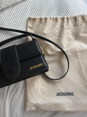 Svart Jacquemus axelremväska - Snygg svart handväska från Jacquemus i slätt läder med guldfärgad logga och detaljer. Väskan har ett lock framtill och en smal axelrem. Perfekt för dig som gillar stilrena accessoarer med exklusiv känsla.