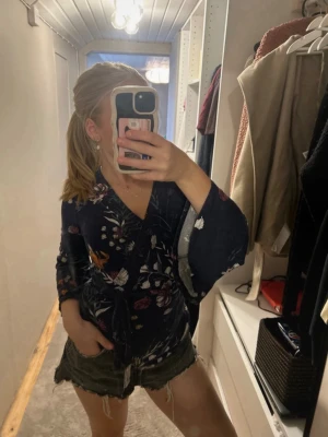 Mörkblå omlottblus med blommor - Snygg mörkblå omlottblus med blommigt mönster i rosa, vitt och orange. Blusen har vida, långa ärmar och knyts i midjan för en feminin siluett. Perfekt att styla med jeans eller kjol för en trendig look.