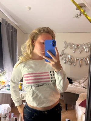 Vit sweatshirt med rosa flaggtryck - Vit sweatshirt med tryck av amerikansk flagga i rosa och svart på bröstet. Tröjan har rund halsringning, långa ärmar och ribbade muddar vid ärmslut och nederkant. Perfekt för en avslappnad och trendig look.