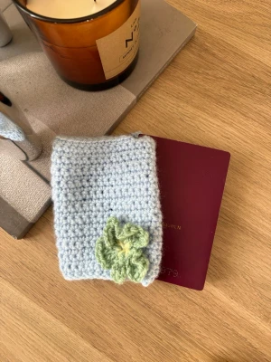 Virkad ljusblå passfodral med blomma - Virkad passfodral i ljusblått garn med en söt ljusgrön blomma på framsidan. Fodralet är mjukt och ger ditt pass en personlig och unik look. Perfekt accessoar för dig som gillar handgjorda detaljer.