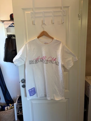 Vit t-shirt med tryck - Säljer en vit t-shirt med trycket 'Health Club 1100 BELMONTE PALACE' i svart och rosa på framsidan. T-shirten har rund hals och korta ärmar. Materialet känns som bomull och passformen är normal. Perfekt till en chill vardagslook.