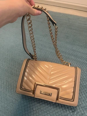 Beige axelväska med kedja River Island - Snygg beige axelväska från River Island med quiltat mönster och glansig finish. Väskan har guldfärgad kedjerem och dekorativ metallspänne framtill.