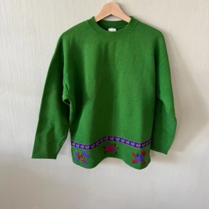 United Colors of Benetton Grön stickad tröja med blommor - Grön stickad tröja med rund hals och långa ärmar. Nedtill finns ett dekorativt band med lila, rosa och röda blommor samt stjärnmönster. Tröjan har en avslappnad passform och ribbade muddar vid halsen. Perfekt för dig som gillar färgglada detaljer.0179