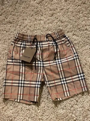  Burberry shorts  - Snygga shorts från Burberry i klassiskt beige rutigt mönster med svarta och röda detaljer. Shortsen har elastisk midja med svart snörning och sidofickor. Perfekta för en trendig och avslappnad stil. Storleken är L men passar S och även M.