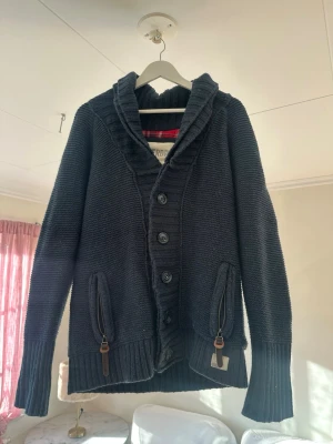 Mörkblå stickad kofta Superdry - Snygg mörkblå stickad kofta från Superdry med grov ribbad krage och stora knappar framtill. Två fickor med dragkedja och läderdetaljer. Insidan har röd- och vitrutigt foder upptill. Perfekt för kyliga dagar.