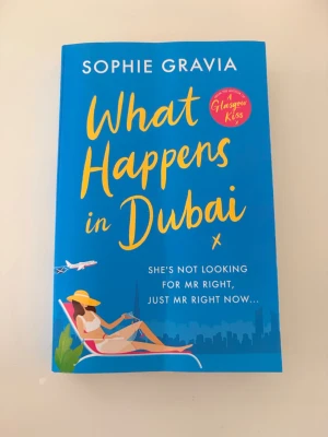 What Happens in Dubai by Sophie Gravia - En färgstark pocketbok med blått omslag och gul text. Omslaget visar en kvinna i solhatt som slappar vid poolen, med ett flygplan i bakgrunden. Boken handlar om Zara Smiths äventyr och flirtar i Dubai, fylld av humor och romantik.