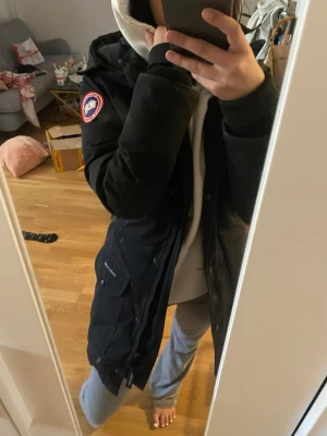 Svart lång dunjacka från Canada Goose - Säljer en svart lång dunjacka från Canada Goose med huva och klassisk logga på ärmen. Jackan har dragkedja och knappar framtill samt stora fickor på sidorna. Perfekt för kalla vinterdagar och riktigt varm.