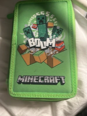 Minecraft pennfodral med pennor - Säljer ett grönt pennfodral med Minecraft-tryck och creepers på framsidan. Inuti finns flera gröna och vita pennor samt en blyertspenna med Minecraft-motiv. Fodralet har dragkedja och är gjort i tyg med tryckt design.