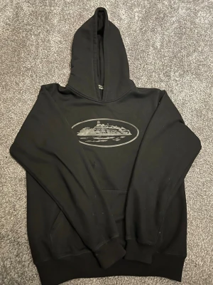 Svart hoodie från corteiz - Säljer en svart hoodie med huva och magficka. På bröstet finns ett tryck av ett fartyg i grått, inramat i en oval. Hoodien har ribbade muddar vid ärmslut och nederkant. Perfekt för dig som gillar enkel men snygg streetwear.