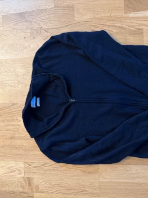 Svart merinoullströja med dragkedja - Stilren svart tröja från These Glory Days i mjuk merinoull. Tröjan har hel dragkedja framtill och en enkel, klassisk design med lång ärm och hög krage. Perfekt för lager-på-lager och passar till det mesta.