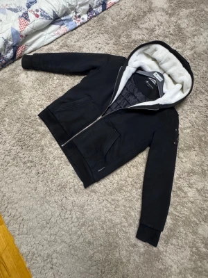 Moose knuckles jacka - Snygg svart hoodie från Mountain Works med dragkedja och stor huva fodrad med vit fuskpäls. Insidan har quiltat mönster och två fickor framtill. Perfekt för dig som vill ha en varm och stilren hoodie med extra mysfaktor.