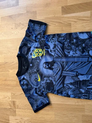 Blå Brasilien t-shirt med grafiskt tryck - Unik blå t-shirt med Brasilien-emblem och grafiskt tryck framtill. T-shirten har korta ärmar, svart rygg och detaljer i gult. Perfekt för dig som gillar fotboll och vill sticka ut med en cool design.