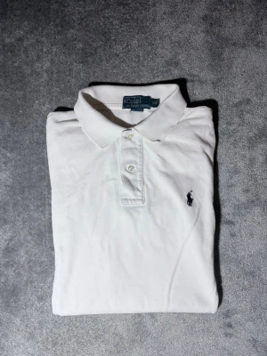 Vit pikétröja från Polo Ralph Lauren - Klassisk vit pikétröja från Polo Ralph Lauren med broderad logga på bröstet. Tröjan har krage, knappar framtill och korta ärmar. Perfekt till en avslappnad men ändå stilren look.