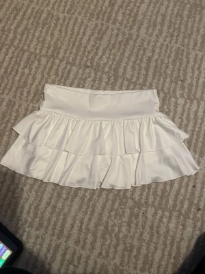 Vit tjol - Supersöt vit skort med dubbla volanger och bred midjeresår. Kjoldesignen ger en flowy känsla och materialet ser ut att vara mjukt och stretchigt, perfekt för en trendig och sportig look. Passar till sneakers eller sandaler för en fräsch outfit. sommar. har short under tjolen är jätte smart.