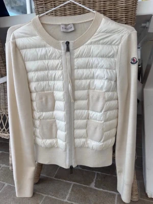 Vit cardiganjacka från Moncler - Stilren vit cardiganjacka från Moncler med quiltad framsida, två framfickor och dragkedja. Ärmar och rygg i mjuk stickad kvalitet, rund halsringning och Moncler-logga på ärmen. Perfekt för lager-på-lager och snygg till jeans.