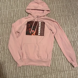 Rosa hoodie med tryck 'THRILLER' - Säljer en ljusrosa hoodie med huva och magficka. På bröstet finns ett stort tryck med en skällande hund och texten 'THRILLER' i vitt. Hoodien har långa ärmar och ribbade muddar. Perfekt för dig som gillar streetwear och coola prints.