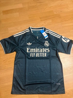 Real madrid 26/27 borta S - Snygg mörkgrön Real Madrid matchtröja från Adidas med vita detaljer och klubbmärke på bröstet. Tröjan har korta ärmar, rund hals och klassiska Adidas-ränder på axlarna. Tillverkad i lätt och ventilerande material, perfekt för fotboll eller träning.