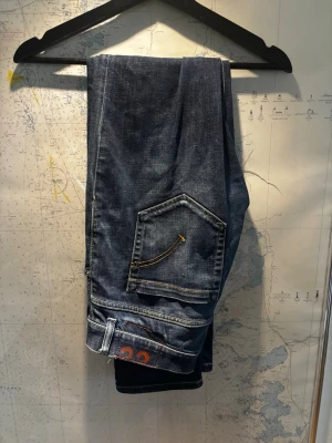 Dondup George  - Säljer ett par blå dondup jeans med raka ben och kontrastsömmar. Jeansen har en unik fickdesign med snedställd ficka och orange detaljsöm. Materialet är slitstark denim i 100% bomull. Perfekta för en avslappnad och stilren look.