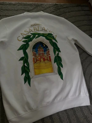 Vit hoodie med Casablanca-tryck - Vit hoodie med färgglatt tryck på ryggen som föreställer en portal med gröna blad och en byggnad under natthimmel. Texten 'Avenida Casablanca' ovanför motivet. Mindre tryck framtill med samma tema. Långärmad och i mjukt material.