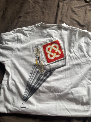 Vit Casablanca t-shirt med tryck - Vit t-shirt från Casablanca med unikt grafiskt tryck på både fram- och baksidan. Trycket har inslag av rött, gult och svart och visar en rulle med Casablanca-loggan. Klassisk passform och rund hals.