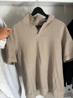 Beige ribbad pikétröja från Zara - Säljer en beige ribbad pikétröja från Zara i storlek M. Tröjan har korta ärmar, öppen krage utan knappar och är gjord i ett mjukt, stretchigt material. Perfekt för en avslappnad och stilren look.
