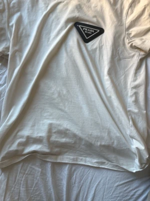 Vit t-shirt med Prada-logga - Säljer en stilren vit t-shirt med en svart Prada-logga på bröstet. T-shirten har klassisk passform och korta ärmar. Perfekt basplagg för en clean look och enkel att matcha med allt.