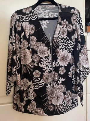 Omlottklänning med dragkedja - Snygg svart klänning med stora beige och vita blommor, omlottmodell och dragkedja framtill med rund metallring. Klänningen har korta ärmar och ett flowy fall, perfekt för dig som gillar statement-mönster. Etiketten saknas. Storleken är uppskattad.