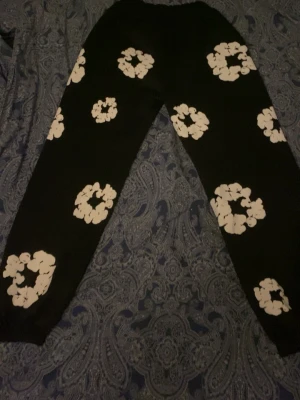 Denim tears byxor - Säljer ett par svarta mjukisbyxor med stora vita blommor tryckta över hela byxorna. Byxorna har resår i midjan och vid bensluten för en skön och avslappnad passform. Perfekta för chill dagar hemma eller när du vill ha en cool look.