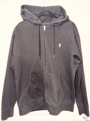 Svart zip hoodie från Polo Ralph Lauren - Klassisk svart hoodie med dragkedja från Polo Ralph Lauren. Hoodien har huva med dragsko, två stora fickor framtill och den ikoniska vita logon broderad på bröstet. Tillverkad i mjukt bomullsmaterial som är perfekt för chill dagar.