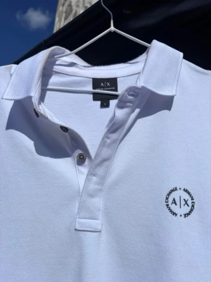 Pikétröja från Armani Exchange -  Använd ett par gånger, vit pikétröja från Armani Exchange med korta ärmar och krage. Tröjan har knappar framtill och en diskret logga på bröstet. Jeans eller shorts.