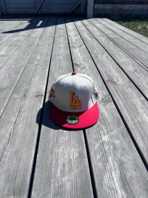 Ny Vit/Röd LA-Keps från New Era - Helt ny fitted vit/röd keps med LA-logga framtill och klassisk rak skärm. Kepsen är från New Era och modellen är 59FIFTY. Perfekt för dig som gillar streetwear och vill ha en stilren accessoar till din outfit. Storlek 55,8 cm