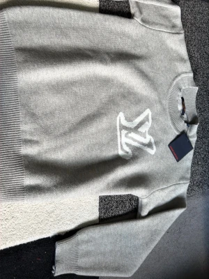 Grå stickad tröja med vit logga - Snygg grå stickad tröja med stor vit logga framtill. Tröjan har rund halsringning och ribbade muddar vid ärmslut och nederkant. Perfekt för dig som gillar enkel men trendig stil. Materialet ser mjukt och bekvämt ut. Storlek S men passar storlek M eftersom den är oversize