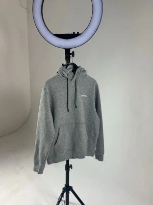 Grå hoodie från Calvin Klein - Snygg grå hoodie från Calvin Klein med klassisk huva och dragsnören. Perfekt som golf hoodie  Perfekt för en avslappnad och trendig look. 