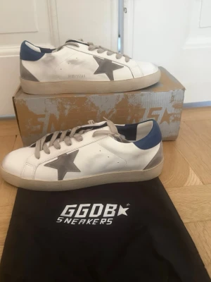 Vita GGDB sneakers med grå stjärna - Säljer ett par vita Golden Goose sneakers med grå stjärna på sidan, beige sula och blå detalj vid hälen. Skorna har klassisk låg profil och är tillverkade i läder med snygga kontrastdetaljer. Perfekta för dig som gillar streetstyle och vill sticka ut.