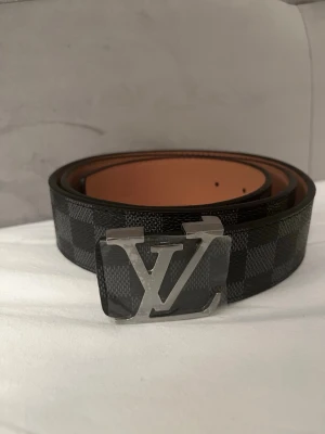 Svart rutigt Bälte - Snyggt svart bälte med diskret rutigt mönster och en stilren logga som spänne. Bältet har en klassisk design och passar perfekt till jeans eller kostymbyxor. Insidan är ljusbrun vilket ger en snygg kontrast.