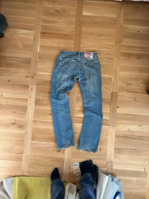 True religion jeans - Säljer ett par blå jeans med flare passform och slitna detaljer vid bensluten. Jeansen har klassiska bakfickor med dekorativa sömmar och en patch bak i midjan. Perfekta för en avslappnad och trendig look.
