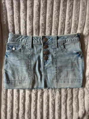 Ljusblå jeanskjol från True Religion - Säljer en ljusblå jeanskjol från True Religion med fyra stora knappar framtill, fickor både fram och bak samt rå kant nedtill. Klassisk denimlook med snygga detaljer och bälteshällor. Perfekt till en avslappnad och trendig stil. Liten fläck som säkert går bort i tvätten💕