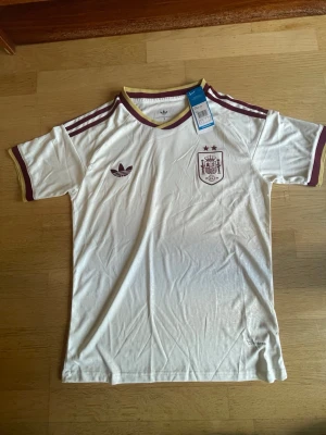 Spanien tröja vm 2026 - Säljer en vit Adidas Spanien tröja med vinröda detaljer och guldkant vid kragen. Tröjan har Adidas-logga och Spaniens landslagsemblem på bröstet samt klassiska ränder på axlarna. Perfekt för dig som gillar fotboll och snygg sportstil. Pris kan diskuteras 