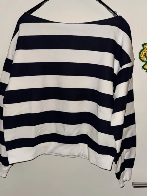 Randig marinblå och vit tröja med båtringning - Säljer en oversized tröja med breda marinblåa och vita horisontella ränder. Tröjan har båtringning och långa ärmar med ribbade muddar. Perfekt för en avslappnad och trendig look.