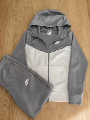 Nike grå och vit tracksuit - Snygg och sportig tracksuit från Nike i grått och vitt. Setet består av en zip-hoodie med fickor och matchande byxor med resår i midjan. Perfekt för träning eller vardag! Båda delarna har Nike-logga broderad i vitt.