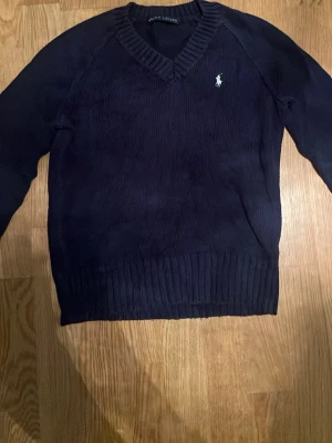 Mörkblå stickad tröja Polo Ralph Lauren - Mörkblå stickad tröja från Polo Ralph Lauren med v-ringning och klassisk vit logga broderad på bröstet. Tröjan har långa ärmar och breda ribbade muddar vid ärmslut och nederkant. Perfekt för lager på lager och stilren look.