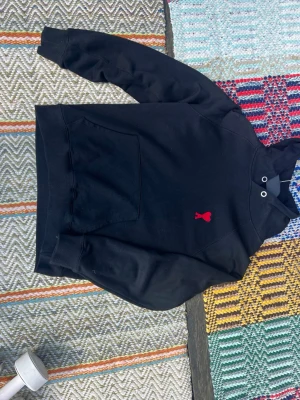 Svart hoodie från Ami Paris - Svart hoodie från Ami Paris med röd broderad logga i form av ett hjärta på bröstet. Klassisk känguruficka framtill och huva med metallöljetter. Tillverkad i mjukt bomullsmaterial, perfekt för en avslappnad och trendig look.