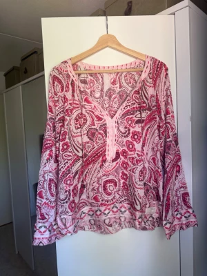 Red and White Paisley Blouse - Lätt blus med lång ärm och ett rödvitt paisleymönster. Blusen har V-ringning med en kort knappslå samt lösa, luftiga ärmar. Tyget är halvtransparent, vilket gör den lämplig att bära i lager-på-lager-stil eller som strandplagg. Nederkanten och ärmsluten pryds av ett dekorativt bårdmönster som kompletterar det övergripande paisleymönstret. Från märket Odd Molly i storlek 3 som enligt deras storlekguide motsvarar en storlek L. Fin men den är tyvärr något tvöttppverkad och kan ha tråddragning.  