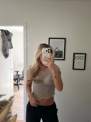 Beige crop top - Säljer en beige croppad topp i spets med korta ärmar. Toppen har ett transparent, mönstrat tyg och rund halsringning. Perfekt för dig som gillar trendiga och luftiga plagg.