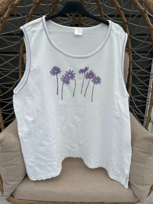 Linnetopp strl. 58/60 - Vit ärmlös linnetopp med lila blommor broderade framtill. Toppen har rund halsringning med lila kantband och en avslappnad passform. Perfekt för varma dagar och enkel att styla med jeans eller shorts.