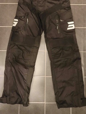 Shot Racing Enduro/Mc byxor - Säljer ett par svarta cargobyxor med vita detaljer och dragkedjeförsedda fickor framtill. Byxorna har förstärkta partier och stretchpaneler vid knäna för extra rörlighet. Materialet är slitstarkt och passar perfekt för en aktiv stil.